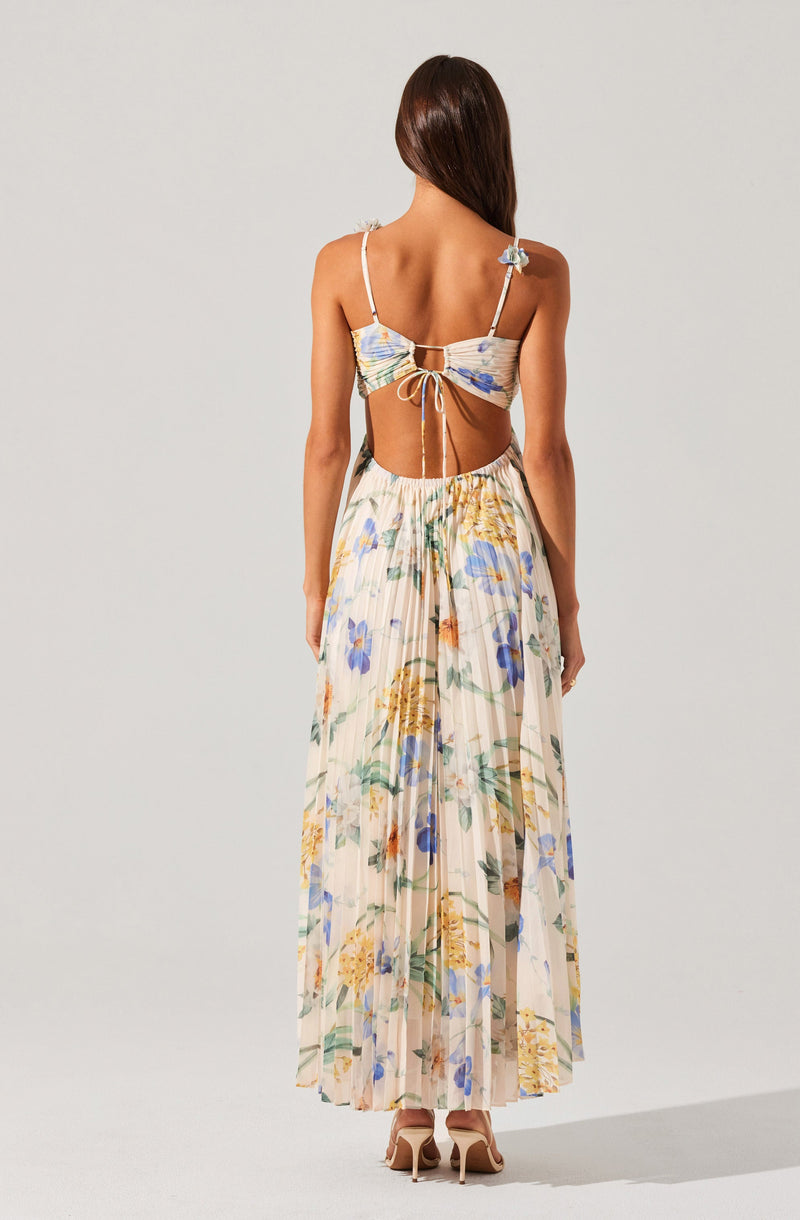 A woman is wearing a light-colored, floral two-piece outfit. The top has thin spaghetti straps and features a small bow at the back, with a cut-out revealing her lower back. The long, high-waisted skirt is pleated with a flowy silhouette, decorated with a multicolored floral pattern including blue, yellow, and green flowers. She is also wearing nude-colored high heels.