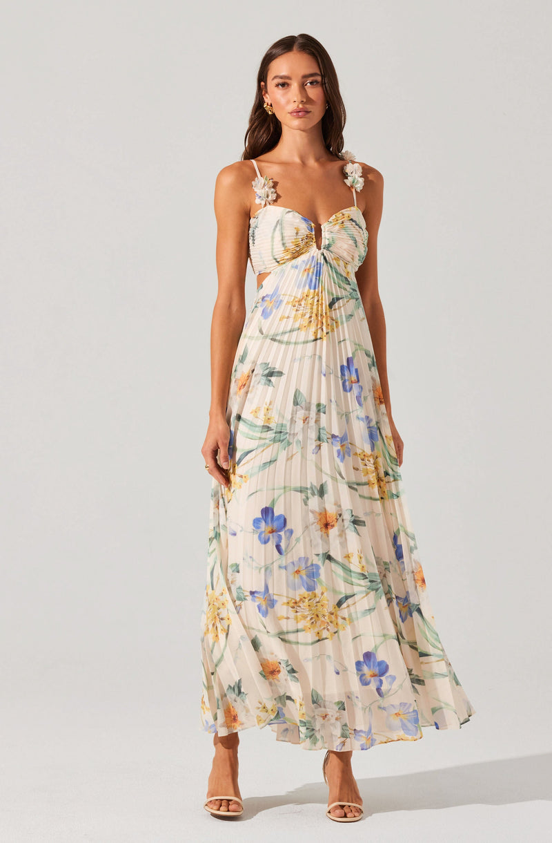A woman is wearing a long, flowing, sleeveless sundress with spaghetti straps adorned with decorative flower appliqués on the shoulders. The dress features a sweetheart neckline with a small keyhole opening and a gathered, ruched bust. The fabric has a light, airy quality with a floral pattern in shades of blue, yellow, and orange on a cream background, with subtle vertical stripes. She is also wearing open-toed high-heeled sandals.