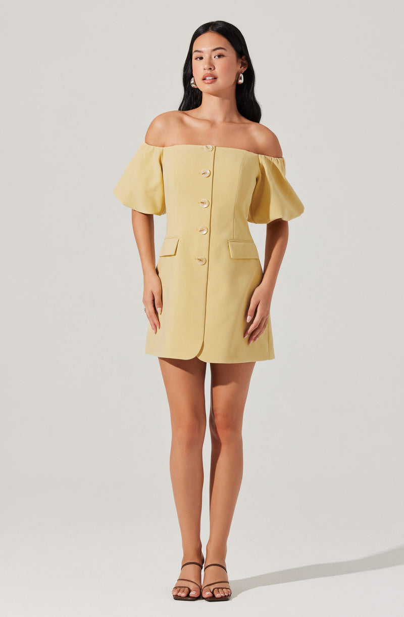 A woman is wearing a light yellow, off-the-shoulder dress with short, puffed sleeves. The dress features large buttons down the front and has two small pocket flaps at the hips. She is also wearing strappy, open-toe sandals and statement earrings.