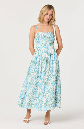 A woman is wearing a sleeveless, ankle-length dress with thin spaghetti straps. The dress features a gathered bust and a waistband, with a floral pattern in shades of light blue, green, and white. She has shoulder-length blond hair and is carrying a small white shoulder bag. She is standing against a plain light background, with one hand behind her and the other holding the strap of her bag. She is wearing white open-toe heels.