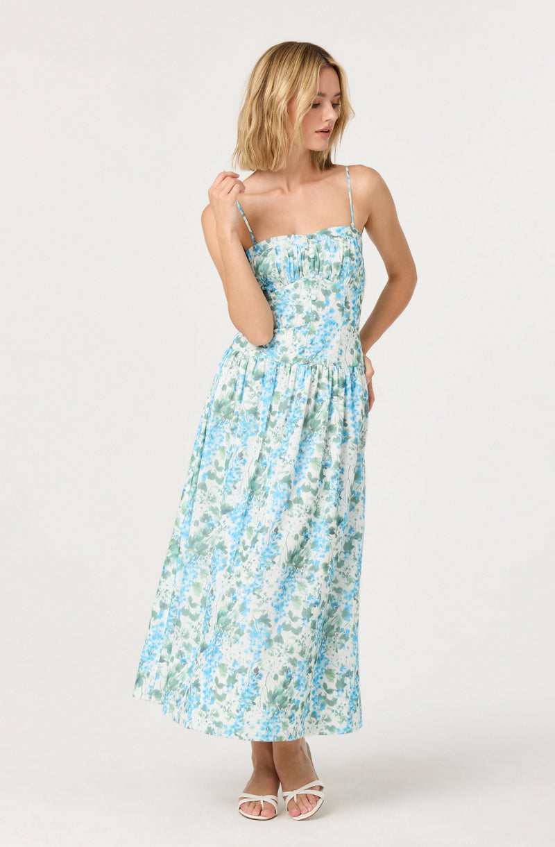 A woman is wearing a sleeveless, ankle-length dress with thin spaghetti straps. The dress features a fitted bodice with a smocked design and a square neckline, and the fabric has a floral pattern in shades of blue, green, and white. She is also wearing white open-toe heels.