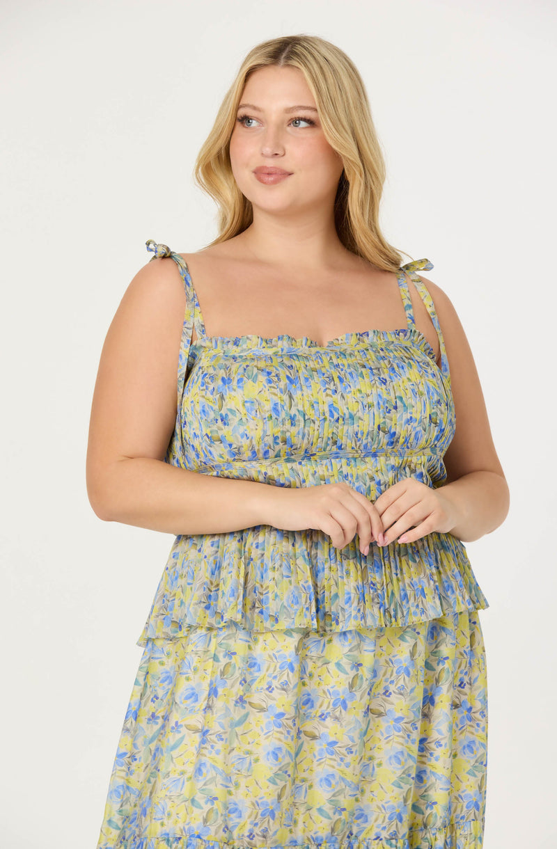 A woman is wearing a sleeveless, floral dress with a yellow and blue pattern. The dress has thin, tied shoulder straps, a ruffled neckline, and a layered, ruffled skirt.