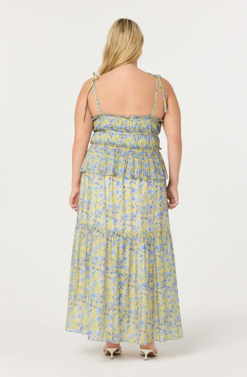 A woman is wearing a sleeveless, layered maxi dress with thin, adjustable spaghetti straps. The dress features a smocked bodice with a ruffled overlay and a floral pattern in shades of blue, yellow, and light green. She is also wearing open-toe, white high-heeled shoes.