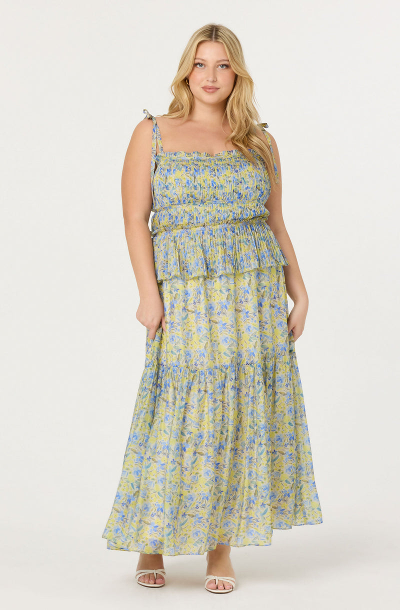 A woman is wearing a sleeveless, floral maxi dress with thin shoulder straps tied into bows. The dress features a smocked bodice and a tiered, flowing skirt, with a light-colored background and blue and yellow floral patterns. She is also wearing open-toe sandals.
