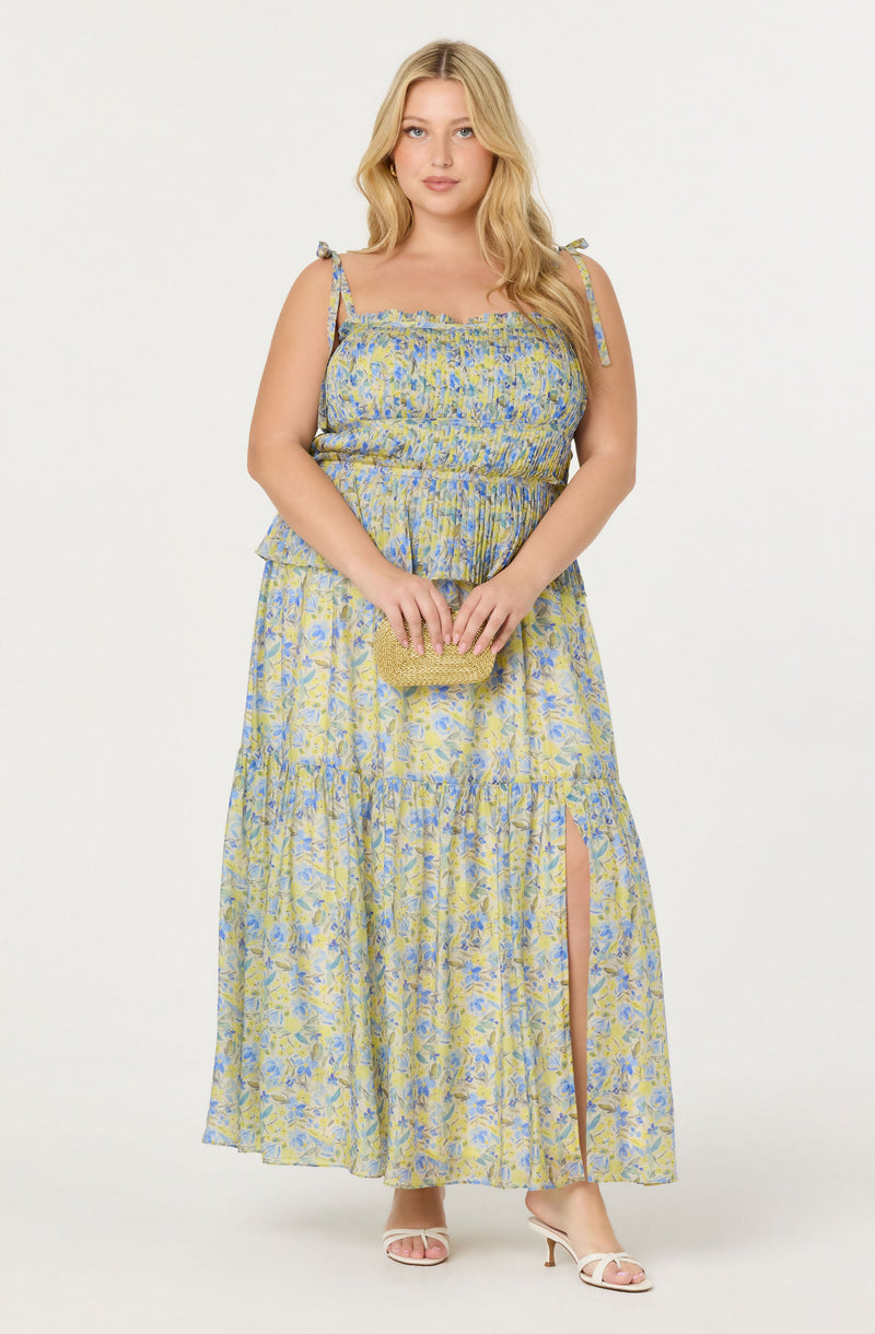 A woman is wearing a sleeveless, maxi dress with a floral pattern in shades of yellow and blue. The dress has thin shoulder straps tied into small bows, a fitted bodice, and a tiered skirt with a slit on one side. She is holding a small, textured, gold clutch purse and is wearing open-toed, white high-heeled sandals.