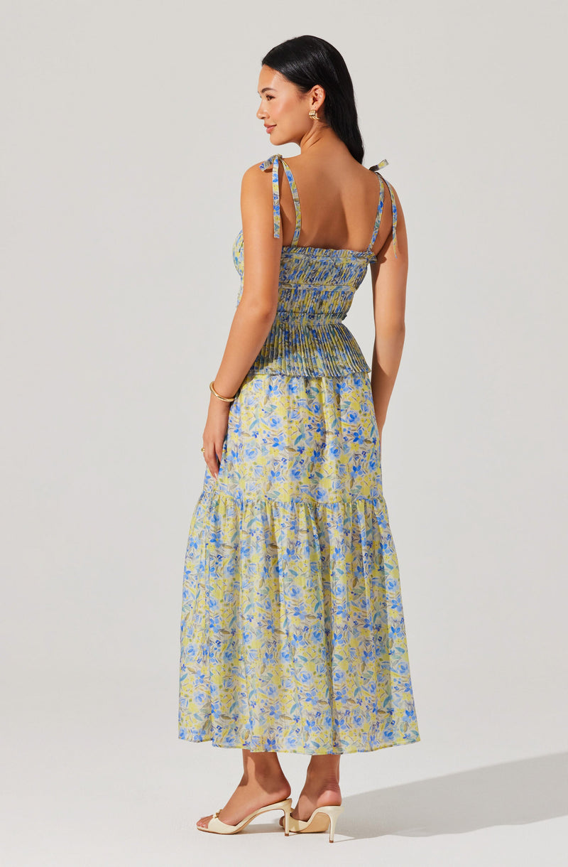 A woman is wearing a sleeveless, floral maxi dress with a fitted, ruched bodice. The dress features adjustable shoulder straps tied into bows at the shoulders. The skirt is flowing and reaches her ankles, with a light yellow and blue floral pattern. She is also wearing open-toed, high-heeled shoes with ankle straps and small earrings. Her hair is long and black, and she has a gold bracelet on her left wrist.
