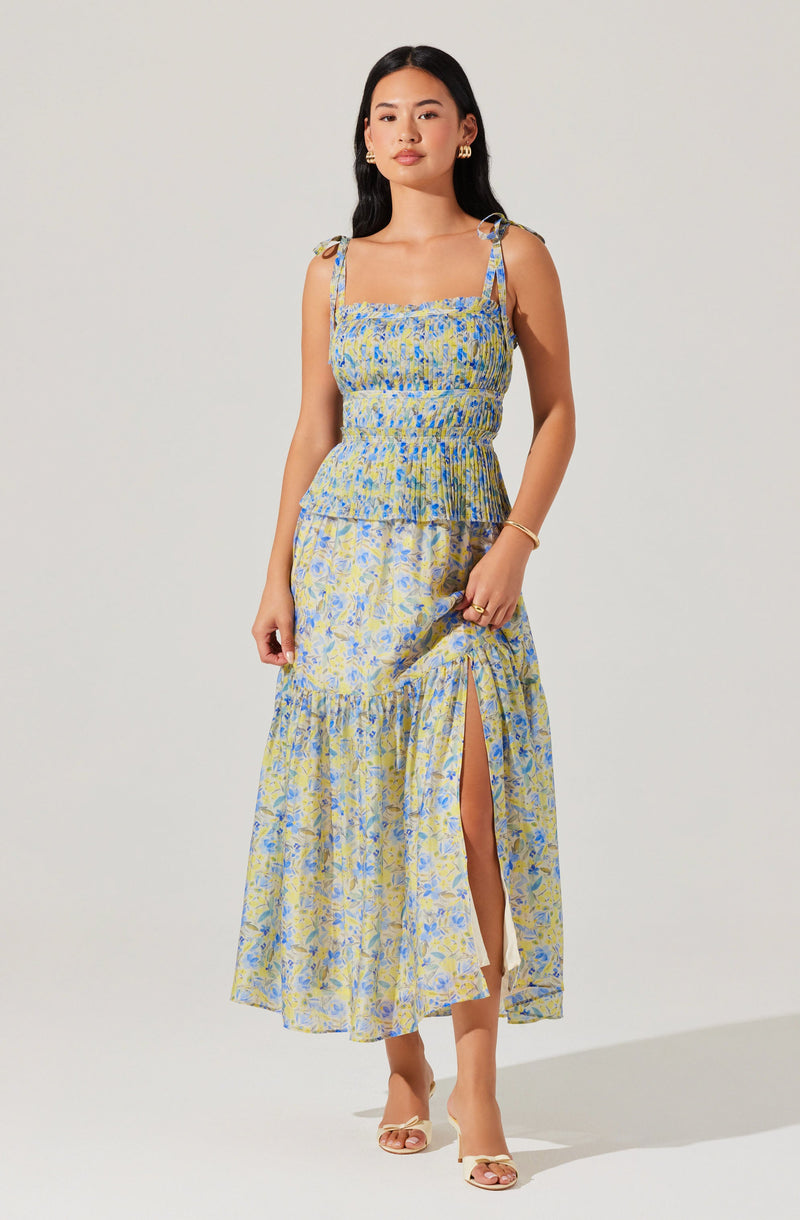 A woman is wearing a sleeveless, ankle-length dress with spaghetti straps tied at the shoulders. The dress features a fitted, smocked bodice and a flowy skirt with a slit up one side. The fabric has a yellow, blue, and green floral pattern. She is accessorized with earrings, a bracelet, and rings, and is wearing open-toe heels.