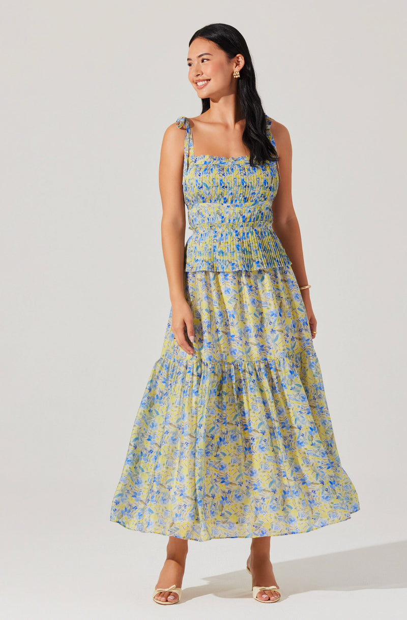 A woman is wearing a sleeveless, square-neck top and a long, flowy skirt. The top has bow-tie straps at the shoulders and features a blue and yellow floral pattern with a smocked, fitted waist. The skirt is made of lightweight, sheer fabric with a similar blue and yellow floral design and has a tiered, ruffled silhouette.