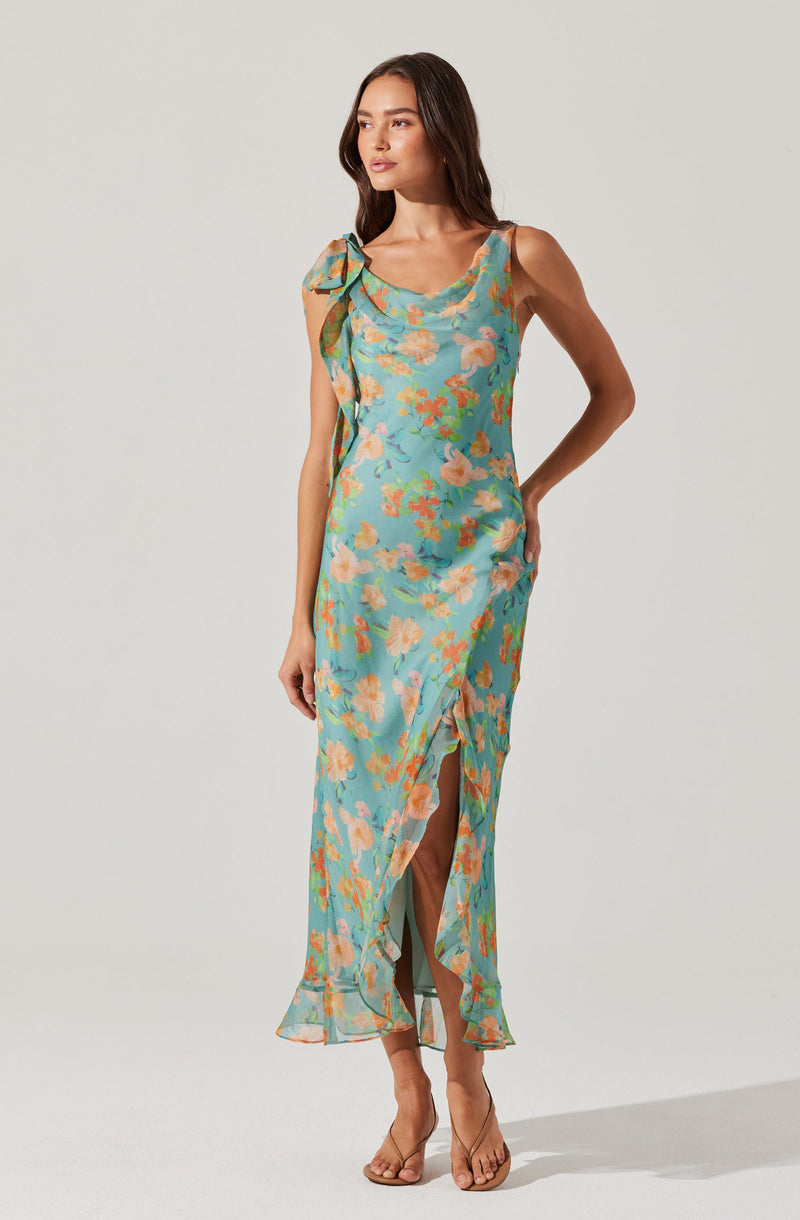 A woman is wearing a sleeveless, ankle-length dress with a floral pattern in shades of blue, peach, and green. The dress features a ruffled neckline, a slit at the front, and a large bow detail on one shoulder. She is also wearing nude-colored, open-toe high heels.