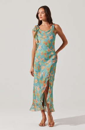 A woman is wearing a sleeveless, ankle-length dress with a floral pattern in shades of blue, peach, and green. The dress features a ruffled neckline, a slit at the front, and a large bow detail on one shoulder. She is also wearing nude-colored, open-toe high heels.