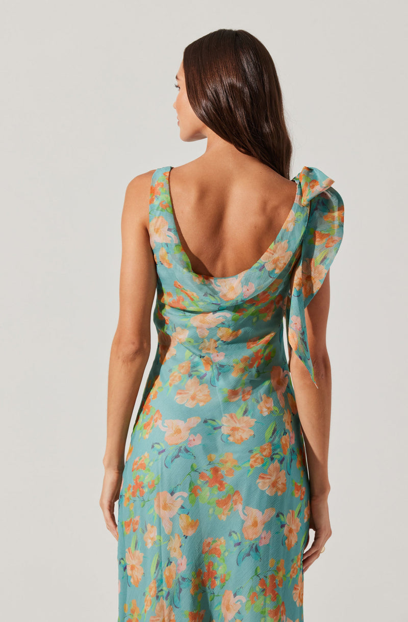 A woman is wearing a sleeveless dress with a floral pattern. The dress has a scoop neckline at the back and features a large, decorative bow or fabric detail on the left shoulder. The fabric is lightweight with a colorful print of orange and beige flowers with green leaves on a light blue background.