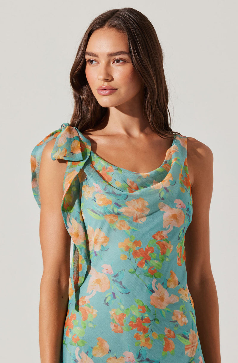 A woman is wearing a sleeveless dress with a round neckline. The dress has a light blue background with a colorful floral pattern featuring shades of peach, orange, pink, green, and purple. The left shoulder has a large, ruffled bow detail made from the same floral fabric. The fabric appears lightweight and slightly sheer.