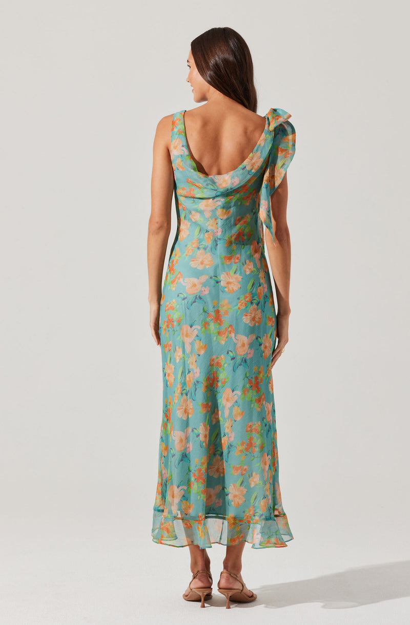 A woman is wearing a sleeveless, ankle-length dress with a floral pattern in shades of peach, green, and pink on a light blue background. The dress features a draped neckline with a large bow on one shoulder, adding a decorative element. The material appears to be lightweight and sheer.