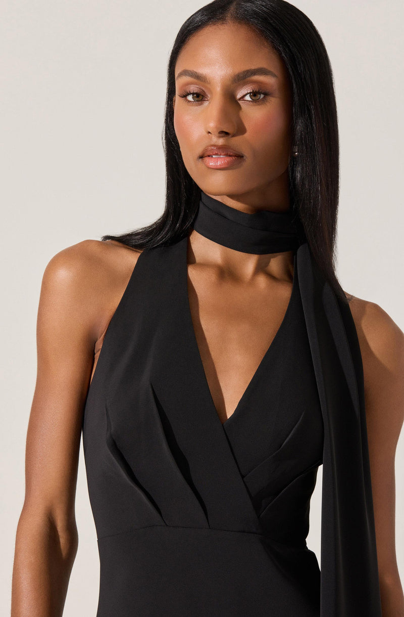 The image shows a woman wearing a sleeveless black dress with a deep V neckline and pleats on the bodice. She has a matching black scarf or necktie wrapped around her neck, hanging loosely over her shoulder. The overall style of the clothing is elegant and sophisticated.