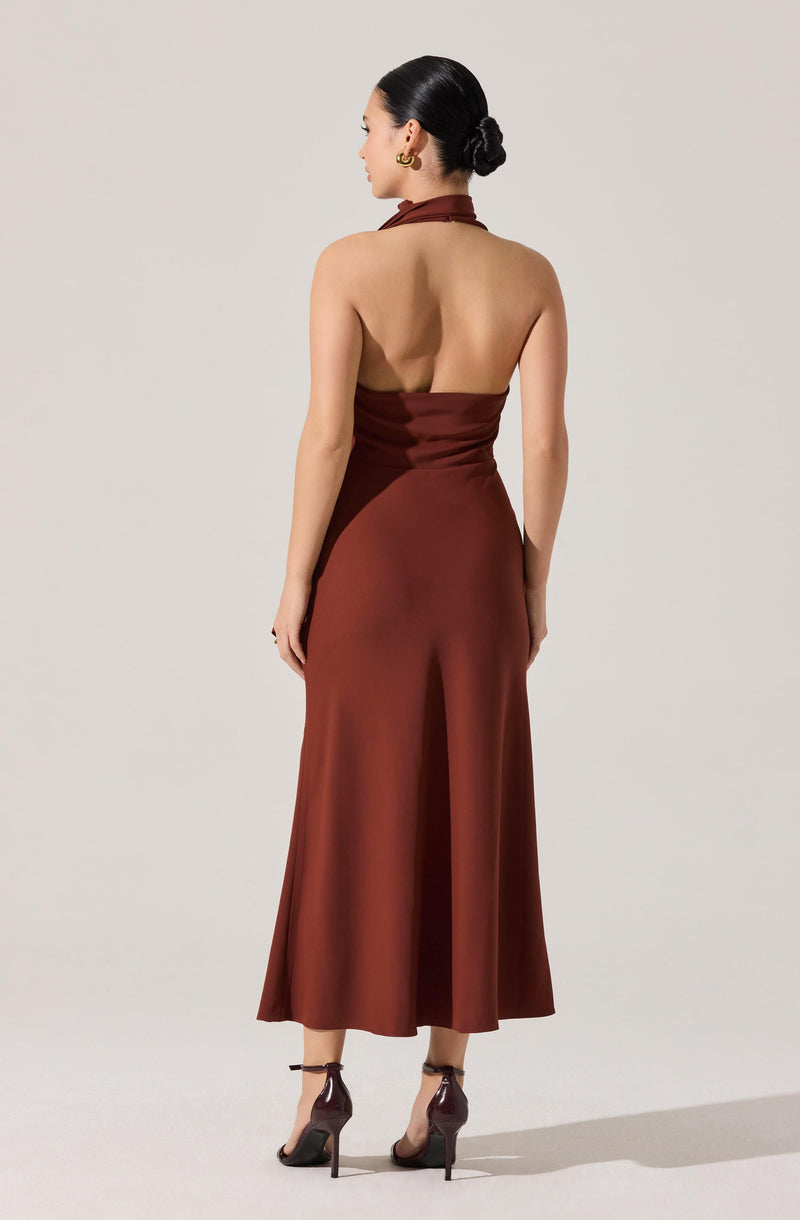 A woman is wearing a sleeveless, halter-neck, satin gown in a rich brown color. The dress features a fitted bodice and a flowing, wide-leg skirt that extends to the floor. She has her hair styled in a neat bun and is accessorized with large, gold hoop earrings and high-heeled sandals with ankle straps.