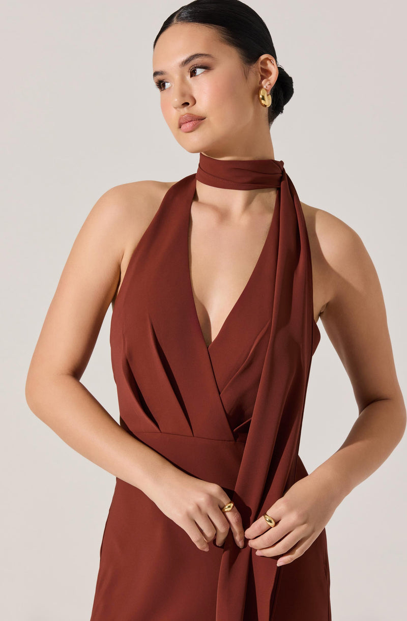 A woman is wearing a sleeveless, deep V-neck dress made of shiny, rust-colored satin fabric. The dress features pleats at the front and a matching fabric scarf tied around her neck, with the ends hanging down. She has dark hair styled in a low bun and is accessorized with gold hoop earrings and gold rings.