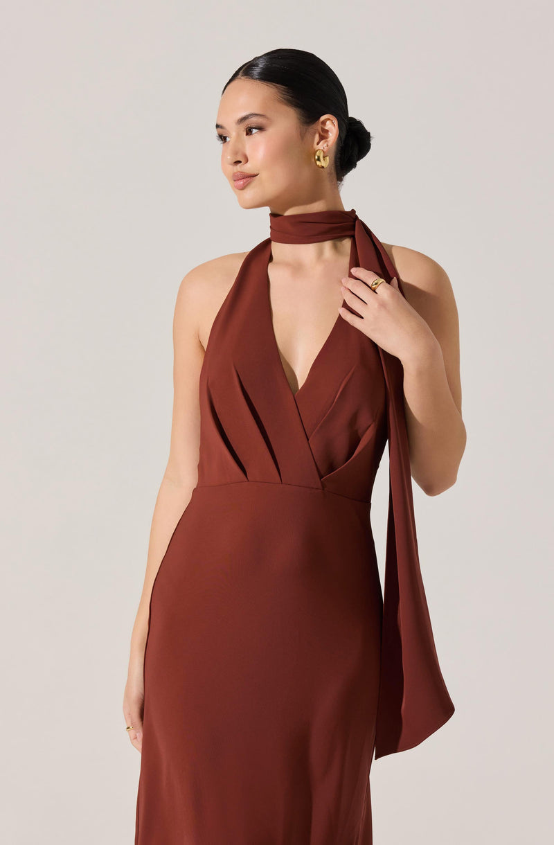 A woman is wearing a sleeveless, deep V-neck satin dress in a rich rust color. The dress features pleats at the bust area and a fitted waist. She has a matching satin scarf tied around her neck, draping down her shoulder. She accessorizes with chunky gold earrings and a ring on her right hand.