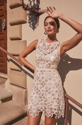A woman wearing a sleeveless, knee-length lace dress with a floral pattern. The dress features intricate cut-out floral designs with circular and petal shapes, and the hemline has a scalloped lace edge. She accessorizes with large hoop earrings and has her left hand raised above her forehead, shading her eyes from the sun. The background includes a beige stucco wall and a wooden door.