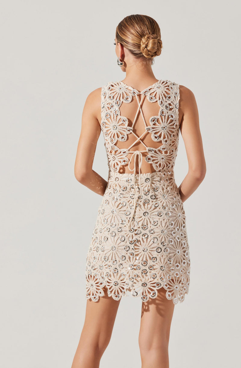 A woman is wearing a sleeveless, knee-length dress made of intricate, white crochet lace with floral and geometric patterns. The back of the dress features a decorative open design with scalloped edges, crisscross lacing, and a tie at the waist.