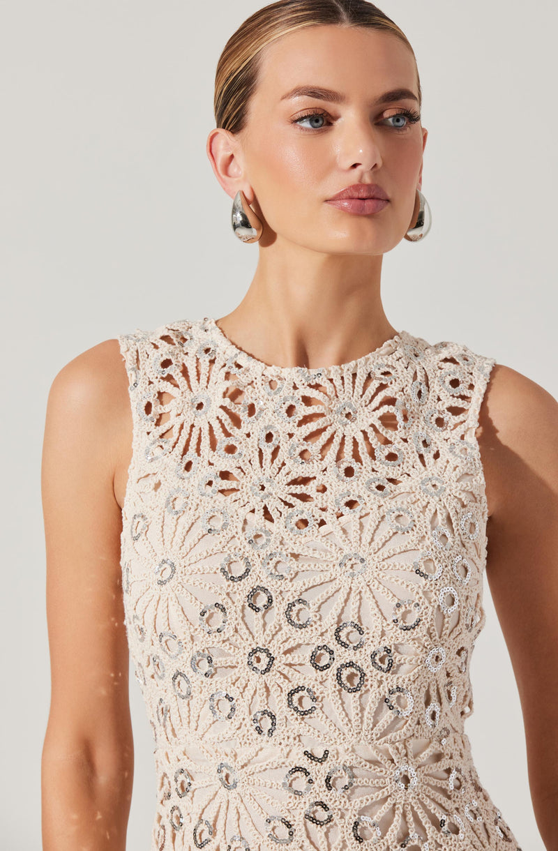 A woman is wearing a sleeveless, cream-colored crochet top featuring a floral pattern with circular and radiating petal designs. The top has a high neckline and intricate openwork details. She accessorizes with large, shiny silver hoop earrings.