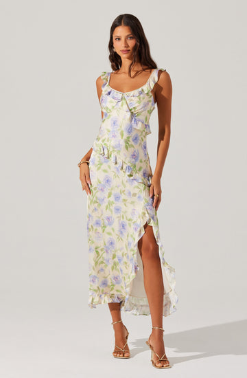 A woman is wearing a sleeveless, ankle-length dress with a floral pattern in shades of purple, green, and white. The dress features ruffled shoulder straps, a ruffled neckline, and a layered, asymmetrical hem with a high slit on one side. She is also wearing open-toed, strappy high-heeled sandals.
