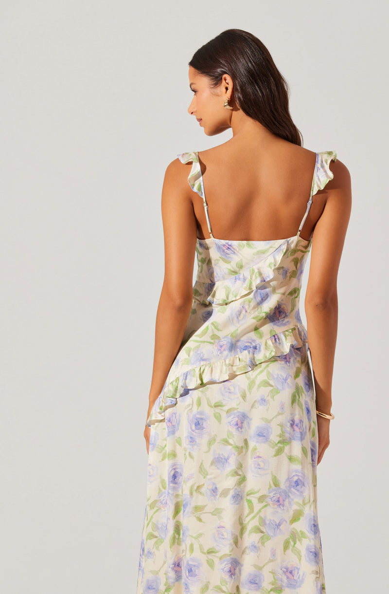 A woman is wearing a sleeveless, ankle-length dress with a floral pattern of light purple and green flowers on a cream background. The dress features ruffled shoulder straps and a ruffle detail across the upper back.