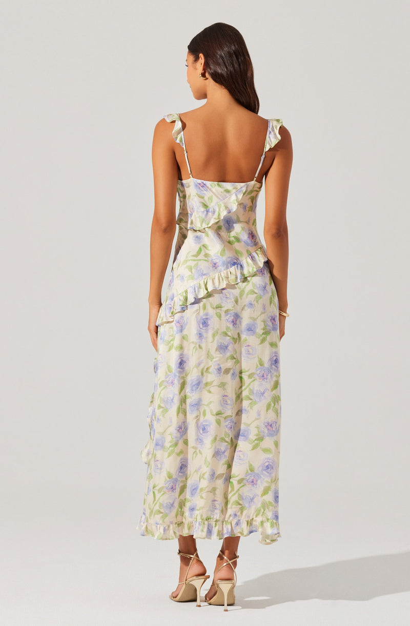 A woman is wearing a sleeveless, ankle-length dress with ruffled shoulder straps and a ruffled overlay at the waist. The dress features a light pastel floral print with shades of purple, green, and cream. She is also wearing beige high-heeled sandals.