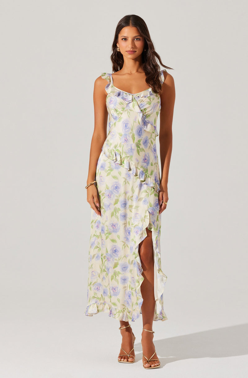 The woman is wearing a sleeveless, ankle-length dress with a light-colored background and a pastel floral print in shades of purple, green, and blue. The dress features ruffle details along the neckline, armholes, and hem, as well as a high slit on one side. She is also wearing nude strappy high-heeled sandals.