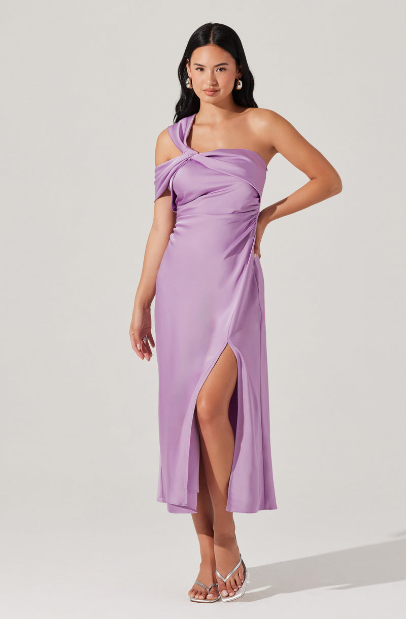 The dress is a lavender-colored, silky, or satin-like gown with an asymmetrical design. It features a one-shoulder strap that drapes across the shoulder and wraps around the chest, creating a gathered, ruched effect on the bodice. The skirt is long and flowing, with a high slit on one side revealing the leg. The woman is wearing silver open-toe heels and statement earrings.