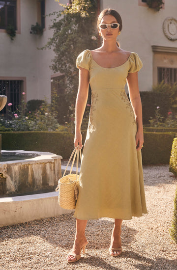 A woman is wearing a light yellow, short-sleeved dress with puffed sleeves and a fitted waist that flares into an A-line skirt. She is accessorizing with white sunglasses, gold earrings, a string bracelet, and beige high-heeled sandals. She is holding a woven straw handbag with a drawstring closure.