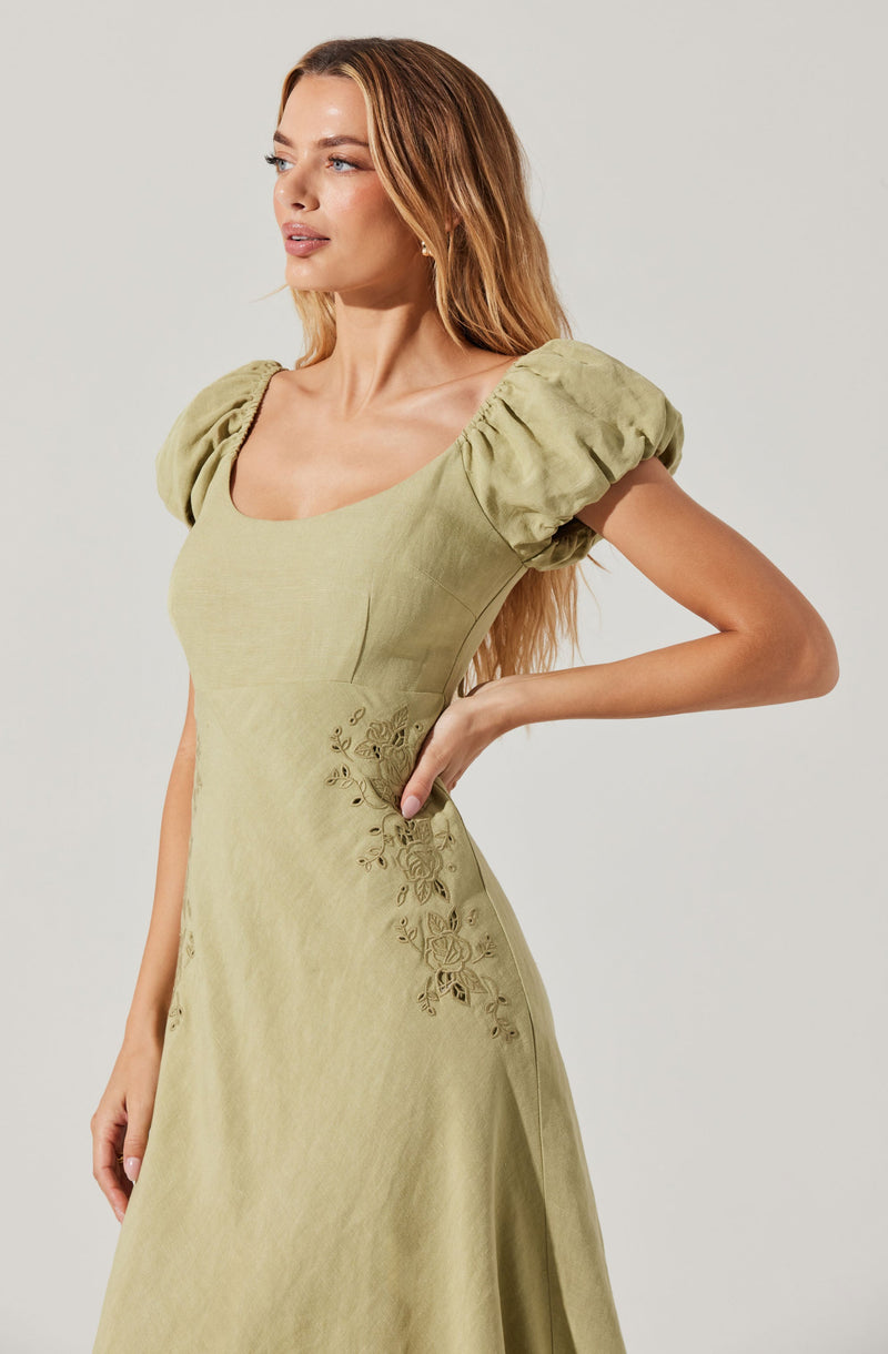 A woman is wearing a light olive-green dress with puffed short sleeves. The dress has a fitted bodice and features delicate floral embroidery on the side. The fabric appears smooth and slightly shiny.