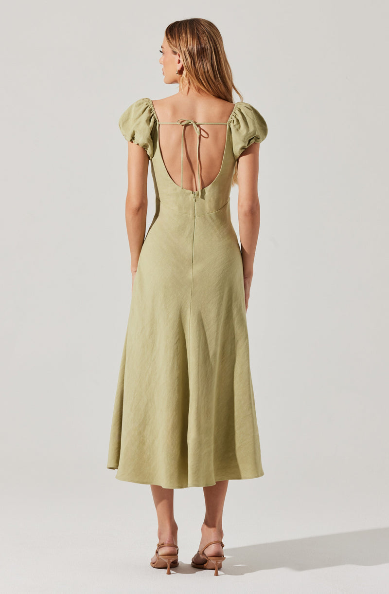 A woman is wearing a light green, ankle-length dress with puffed short sleeves. The dress features a deep, open back with a tie detail at the top and a visible zipper running down the center back. She is also wearing nude-colored high-heeled sandals.