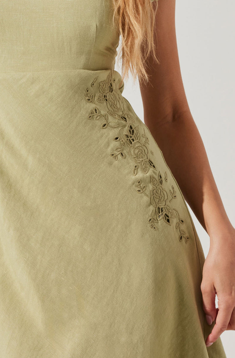 A close-up of a light green dress made of smooth fabric featuring intricate floral embroidery with roses and leaves along the side. The embroidery is detailed with cut-out sections, adding texture to the design. The dress is fitted at the waist, and a portion of the person's arm and hand are visible, with long, wavy hair seen in the background.