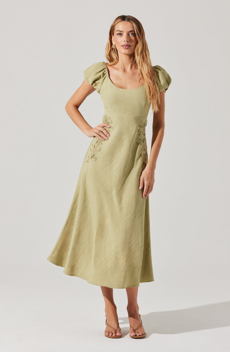 A woman is wearing a long, light green dress with short, puffed sleeves and a wide, rounded neckline. The dress has subtle lace or embroidery details on the left side of the bodice and the waist area. She is also wearing nude high-heeled sandals.