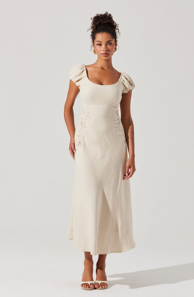 A woman is wearing a long, cream-colored dress with puffed short sleeves and a square neckline. The dress has embroidered or cut-out details on the waist and hips and falls to her ankles. She is also wearing tan heeled sandals.