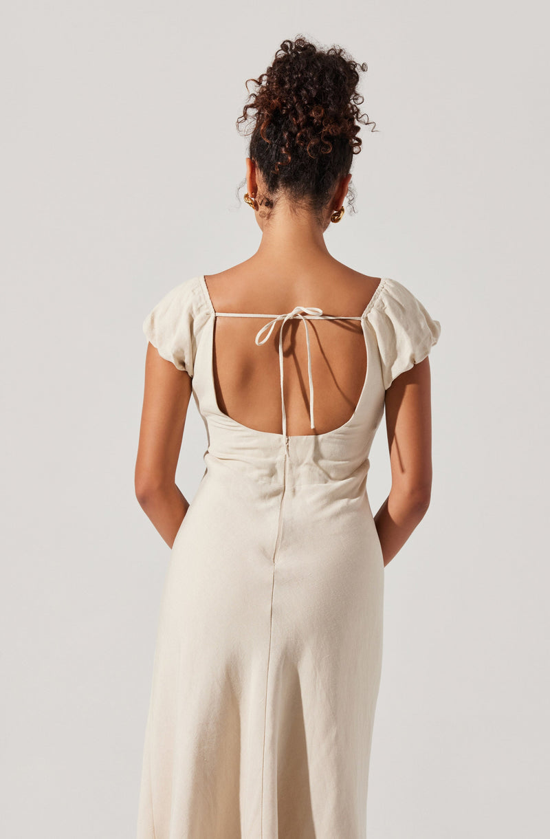 A woman is wearing a cream-colored, satin-like dress with short, puffed sleeves. The dress has a low open back with a thin strap tied in a bow at the top. The material appears smooth and shiny, and the dress extends down past her hips.