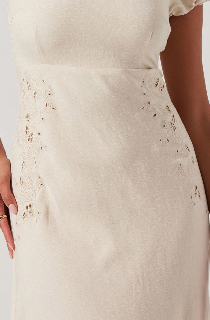 A person is wearing a cream-colored dress with short sleeves. The dress features intricate floral cutout embroidery on the sides, revealing a bit of skin beneath. The fabric appears smooth and slightly shiny.