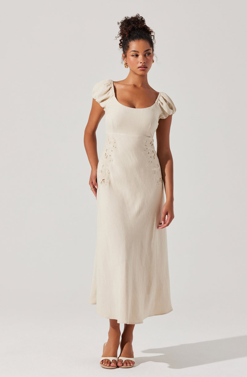 A woman is wearing a long, cream-colored dress with puffed short sleeves and a square neckline. The dress has a fitted waist and features delicate eyelet embroidery on the sides. She is also wearing open-toe nude heels and has her hair styled in a curly bun.