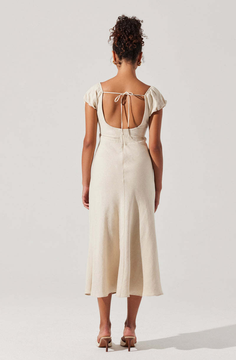 A woman is wearing a cream-colored, sleeveless, ankle-length dress with a fitted waist and a front slit. The dress features a low back with a tie-up detail at the shoulders. She is also wearing a pair of beige high-heeled sandals.