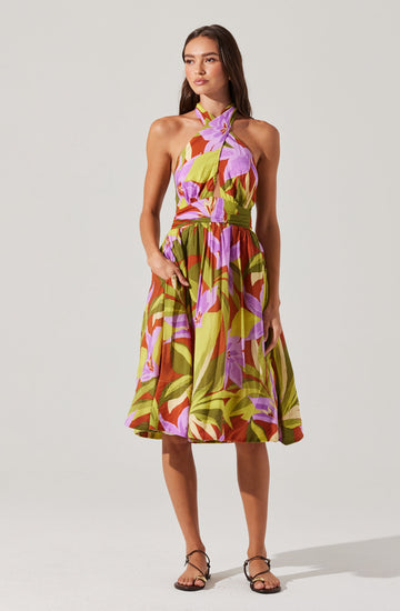 A woman is wearing a sleeveless, halter-neck dress with a colorful, large floral pattern in shades of purple, pink, green, and brown. The dress has a fitted bodice and a full, knee-length skirt.