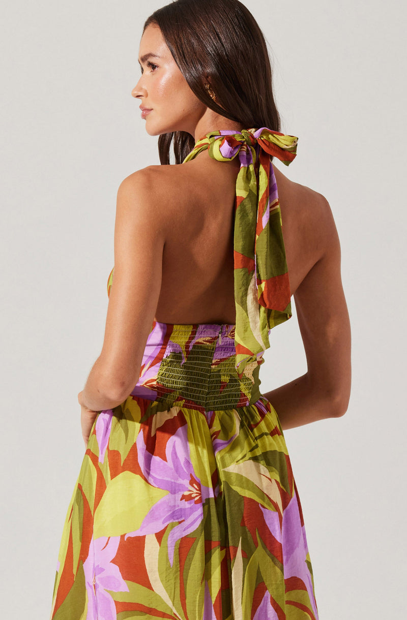 A woman is wearing a colorful, sleeveless, backless dress with a halter tie strap around her neck. The dress features a vibrant floral pattern in shades of purple, pink, orange, and green. The fabric appears lightweight and flowy.