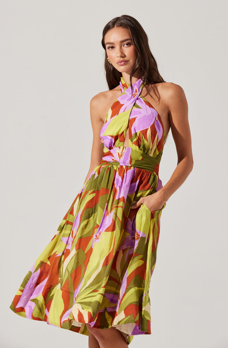 A woman is wearing a sleeveless, knee-length dress with a colorful abstract floral pattern. The dress features a halter neckline and a fitted waistband with a gathered skirt that has large pockets. The fabric appears lightweight and flowy, with a vibrant mix of purple, green, orange, and beige colors.