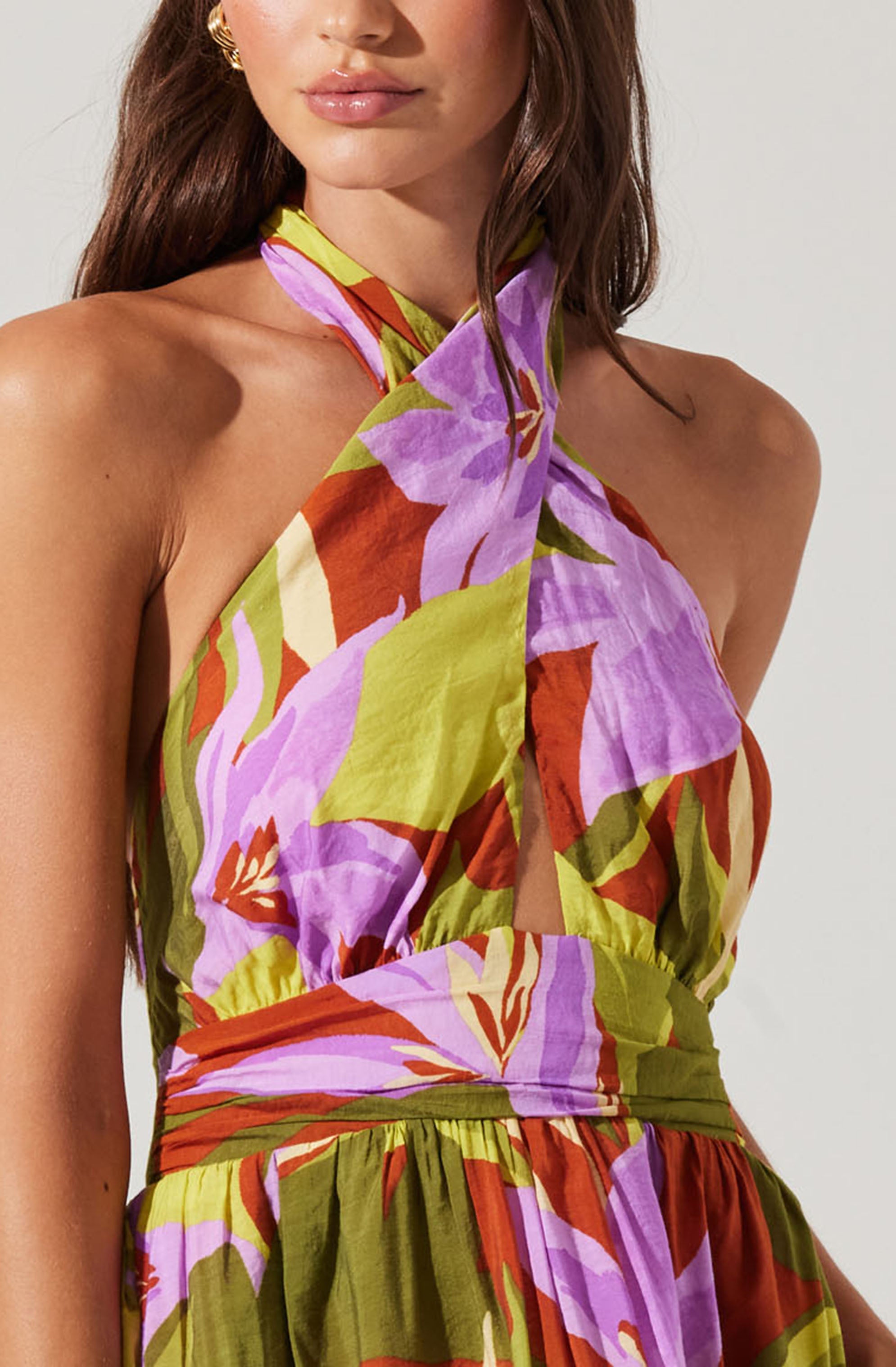 A woman is wearing a sleeveless, halter-neck dress with a vibrant floral pattern in shades of purple, red, and green. The dress features a keyhole opening at the front and a gathered waistline. The fabric appears lightweight and flowy.
