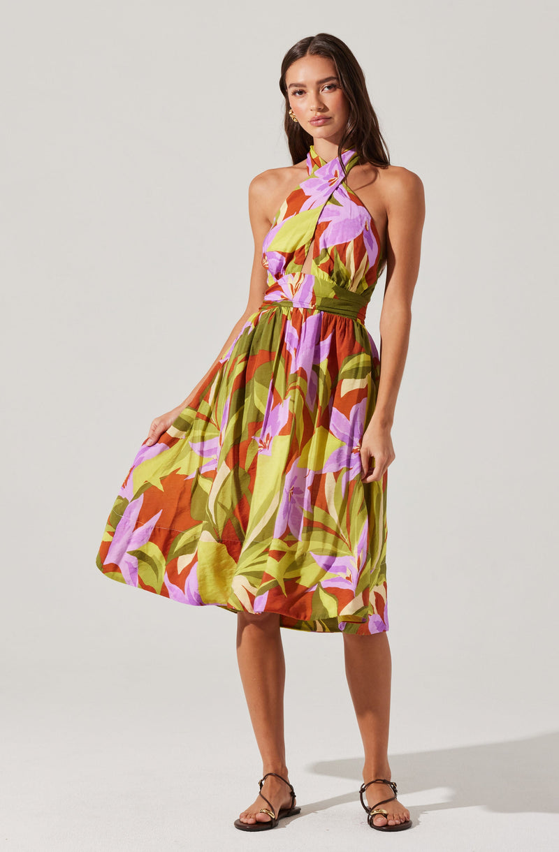 A woman is wearing a sleeveless, halter-neck dress with a colorful, abstract floral pattern in shades of purple, green, yellow, and orange. The dress has a fitted waist and a knee-length, full skirt. She is wearing open-toe, strappy sandals in a dark color.