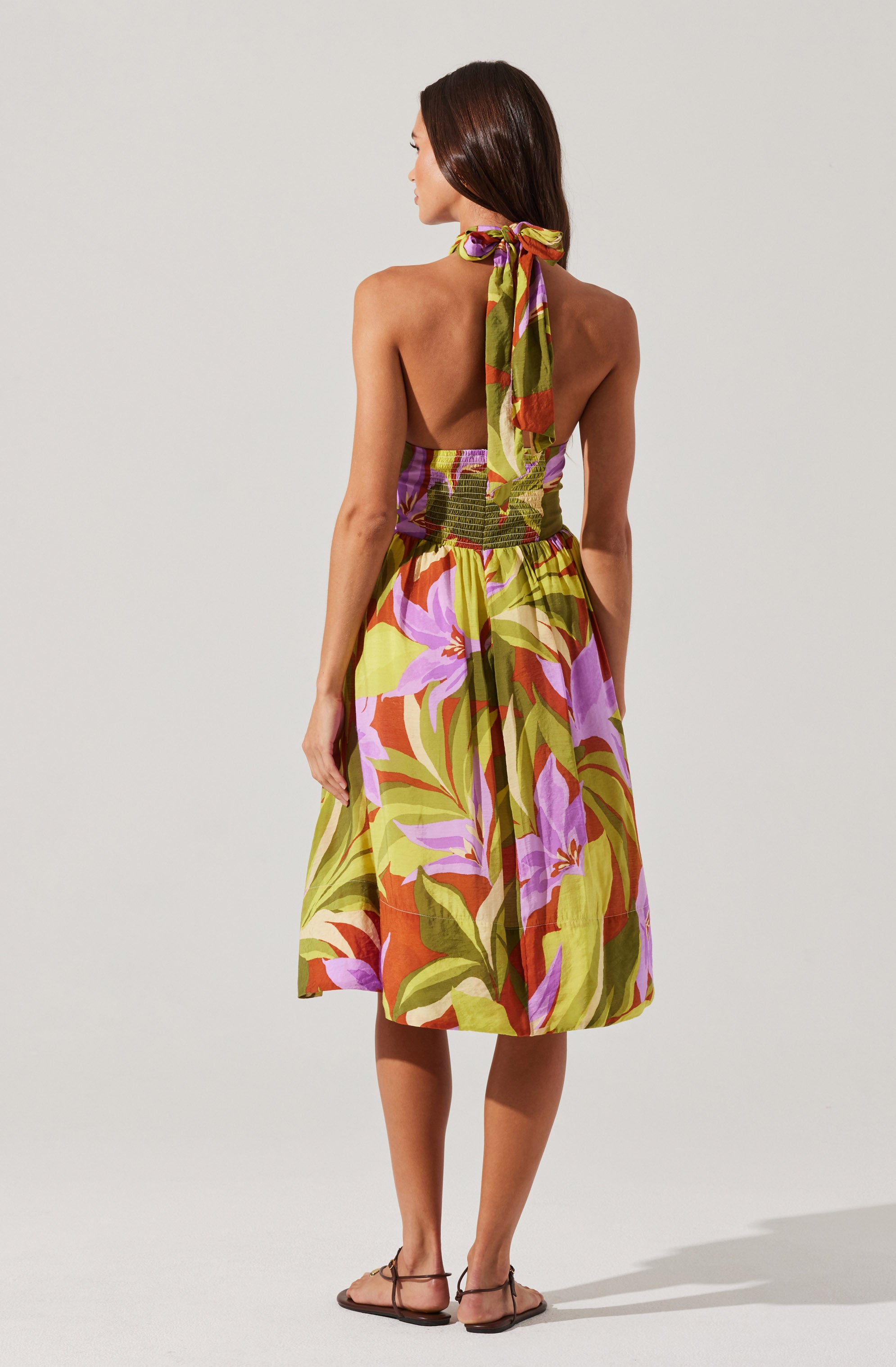 A woman is wearing a strapless, colorful dress with a floral pattern in shades of purple, pink, green, and orange. The dress has a fitted bodice with an elastic waistband and a full, knee-length skirt. She has a matching scarf tied around her neck, which features the same vibrant floral pattern. She is also wearing flat sandals.