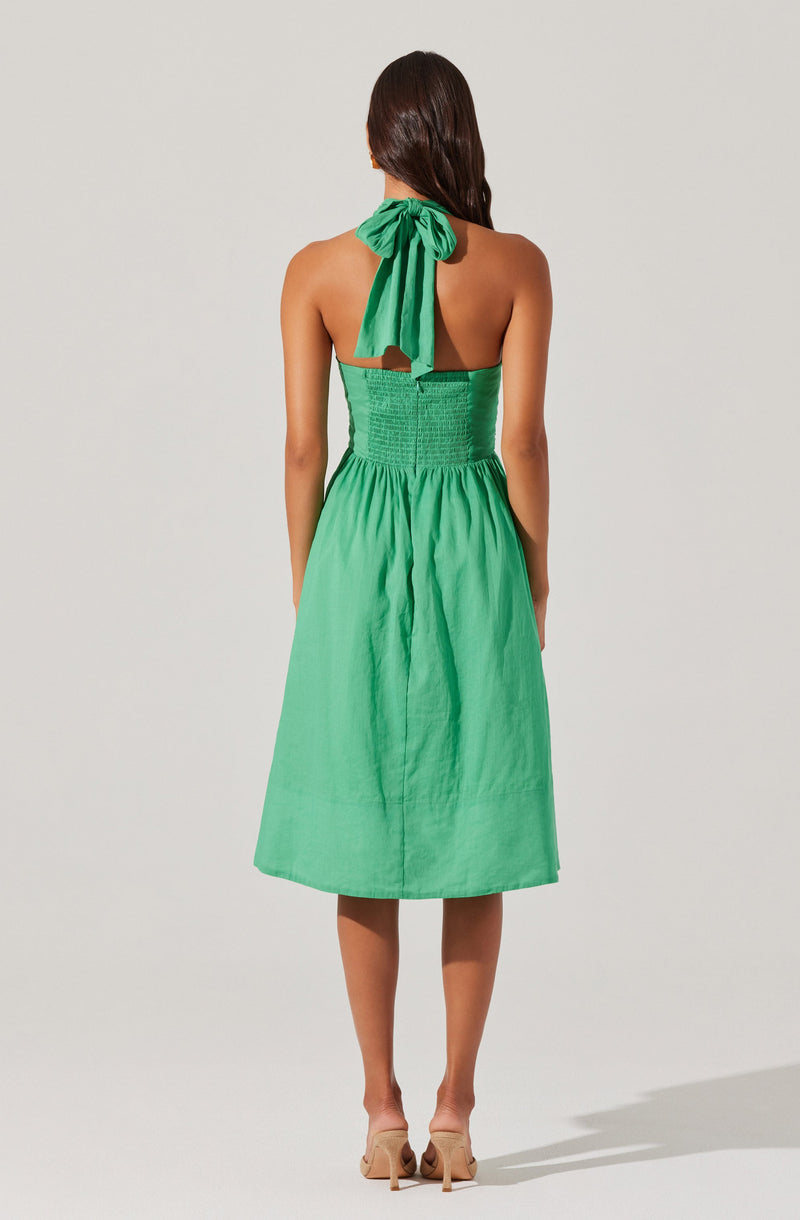 The dress is sleeveless with a halter neckline tied at the neck with a large bow. The bodice is fitted with shirred, textured fabric, and the skirt is flowy and knee-length, made of a lightweight, solid green fabric. The model is wearing nude high-heeled shoes.