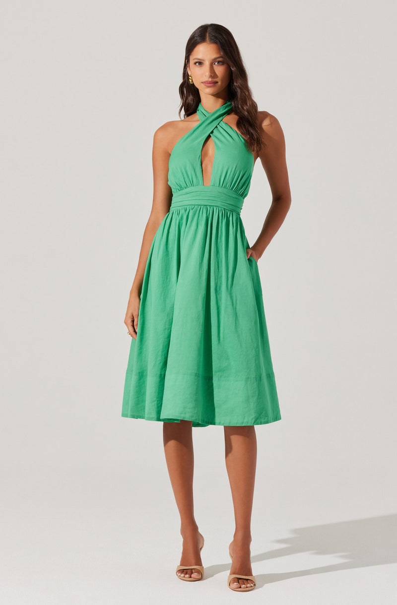 A woman is wearing a sleeveless, halter dress in a bright green color. The dress features a criss-cross halter neckline with a keyhole cutout in the front. It has a fitted waistband and a full, knee-length A-line skirt. The fabric appears lightweight and slightly textured. She is also wearing nude open-toe high heels.