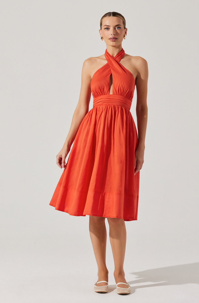 A sleeveless, halter-neck dress with a keyhole opening at the chest, fitted at the waist with a wide waistband, and a full, gathered skirt that falls to knee length. The dress is made of a shiny, satin-like fabric in a bright coral color. The woman is wearing earrings and neutral-colored flats.