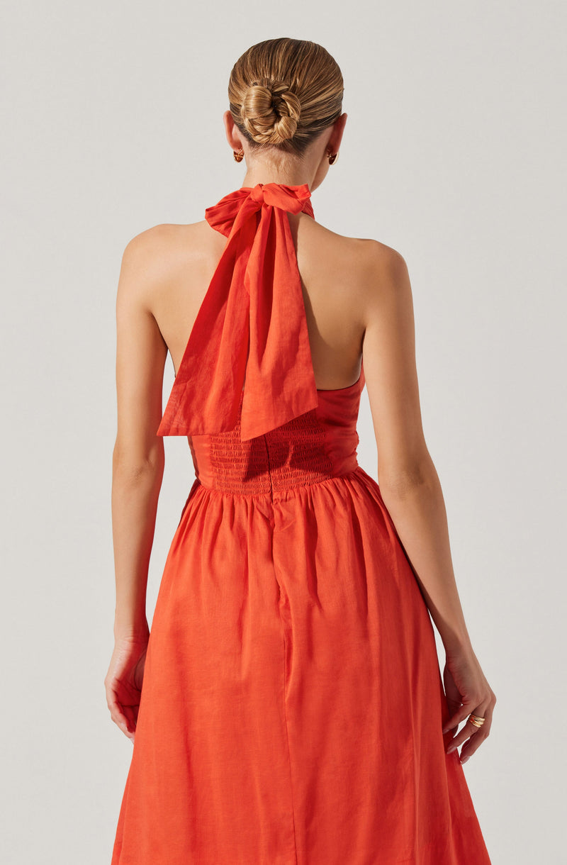The image shows a woman from behind wearing a sleeveless, strapless dress in a coral or burnt orange color. The dress has a fitted, ruched bodice and a gathered, flowing skirt. She has her hair styled in a neat bun and is accessorized with small earrings and a ring. A notable feature is the large, matching orange bow tied around her neck, which drapes down her back.