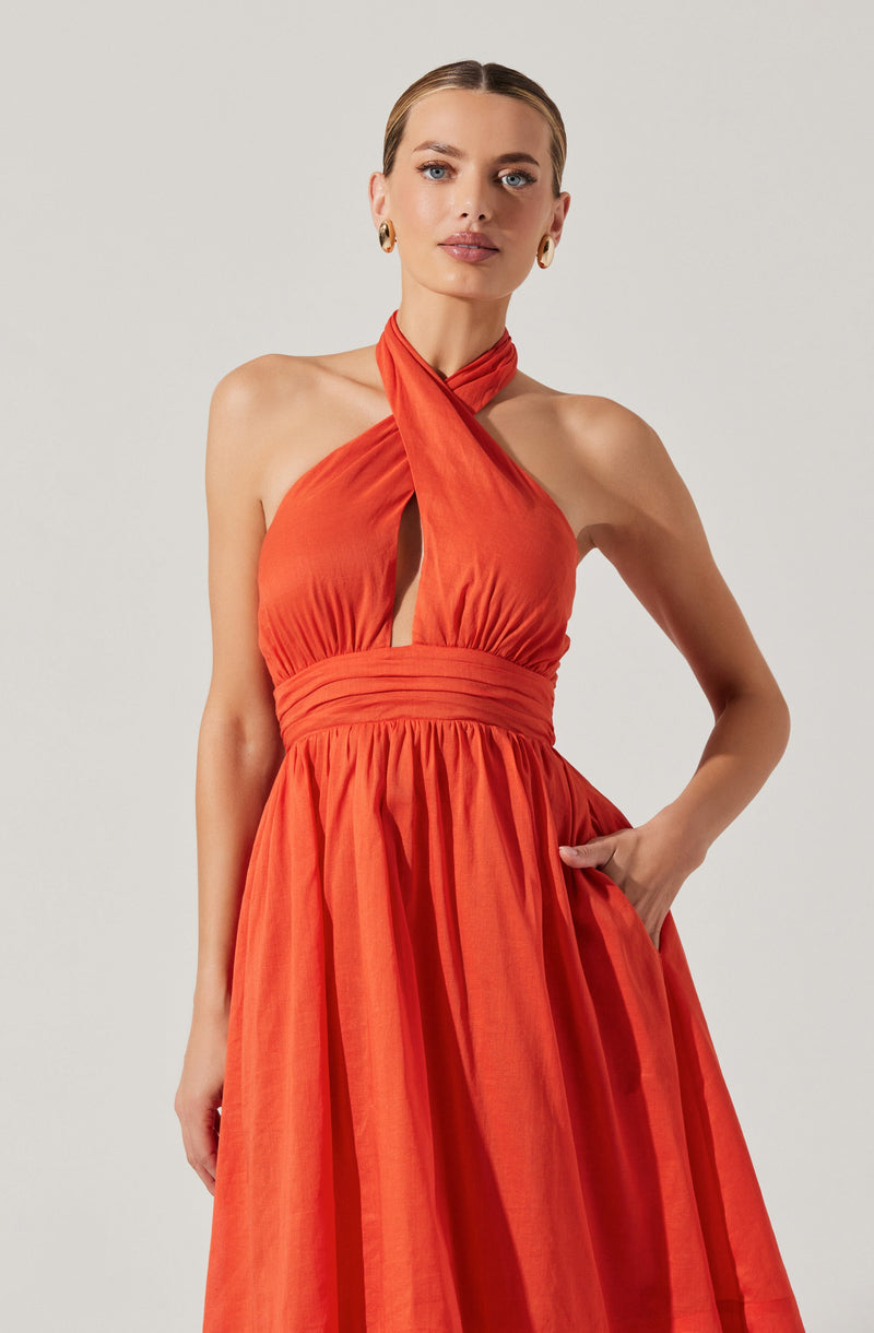 A woman is wearing a sleeveless, vibrant orange halter dress with gathered fabric at the bust and a fitted, ruched waistband. The dress features a high neckline with a knot detail at the neck, creating a keyhole cutout at the front. The skirt is full and pleated, falling to the floor. She accessorizes with gold hoop earrings.